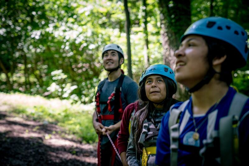 The image shows three people wearing helmets, possibly engaging in an outdoor adventure activity like zip-lining or a ropes course. The person in the foreground is looking to the right, while the other two are behind them on a path in a wooded area. They appear to be wearing safety harnesses and gear, suggesting they are preparing for or participating in a challenging activity.